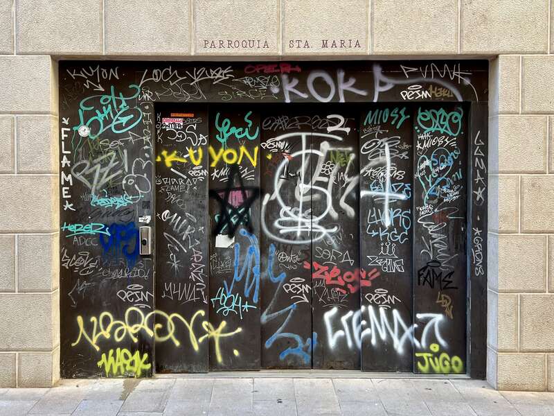 Porta de l’ascensor d’un garatge en un carrer, coberta de grafits de colors. Damunt de la porta, en lletres de motle: “PARROQUIA STA. MARIA”