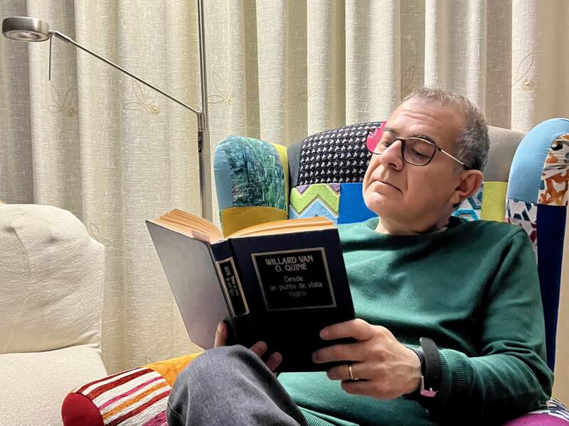 Foto d’un home de mitjana edat amb ulleres, assegut en una butaca, llegint un llibre amb cara d’interès.