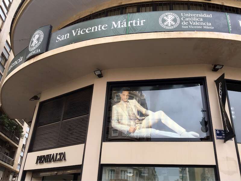 Fotografia d’una façana, amb un rètol de la Universidad Católica de Valencia San Vicente Mártir i, just davall, publicitat d’una botiga de roba amb la imatge d’un model masculí vestit de forma elegant